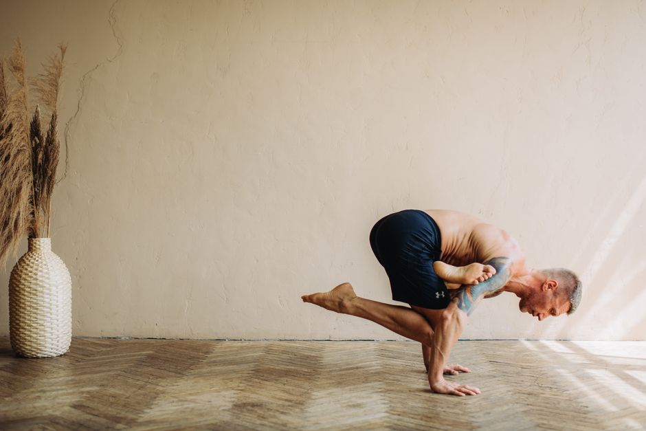 A shirtless man performs a strong crow pose indoors, showcasing flexibility and balance.